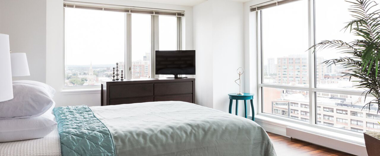 A bed, a dresser and tv in an apartment unit, with large windows overlooking The Fenway neighborhood in Boston.